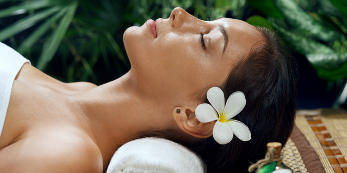 Профиль расслабленной женщины с закрытыми глазами и белым цветком на голове в спа-обстановке.